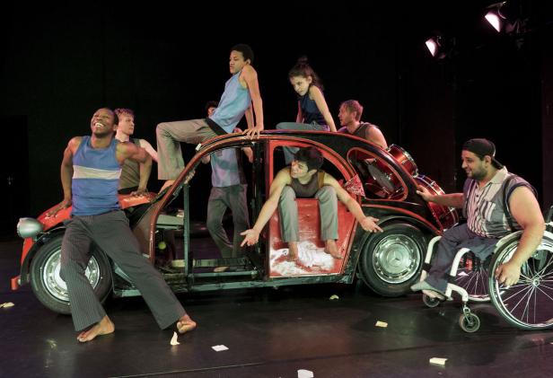 Eine Gruppe von Darstellern interagiert mit einem umgebauten Auto auf einer Bühne.