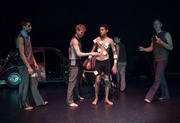 Mehrere barfüßige Darsteller stehen auf einer Bühne, einer mit Haftnotizen beklebt.