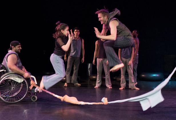 Eine Frau springt über ein Seil, während ein Mann im Rollstuhl zusieht; weitere Personen stehen im Hintergrund.
