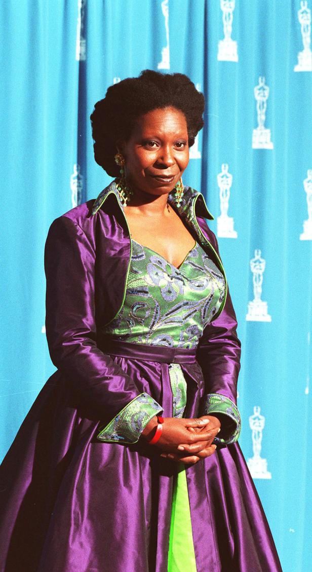Whoopi Goldberg in einem lila-grünen Kleid vor einem blauen Hintergrund mit Oscar-Statuetten.