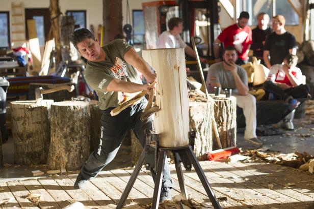 Ein Mann spaltet mit einem Keil Holz, während andere im Hintergrund zusehen.