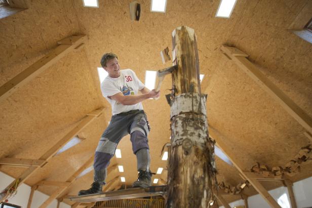 Ein Mann steht auf einem Gerüst und spaltet einen Baumstamm mit einer Axt.