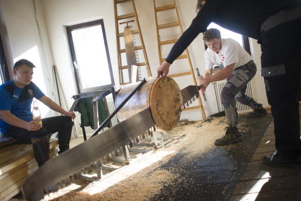 Zwei Männer sägen mit einer großen Schrotsäge einen Baumstamm in einem Innenraum.