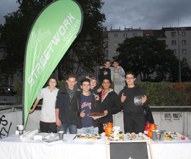Eine Gruppe Jugendlicher steht an einem Stand mit Kuchen und Getränken vor einem Wohnhaus.