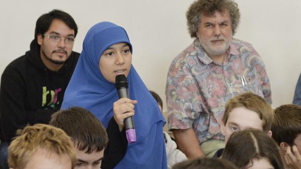 Eine junge Frau mit blauem Hijab spricht in ein Mikrofon vor einer Gruppe von Menschen.
