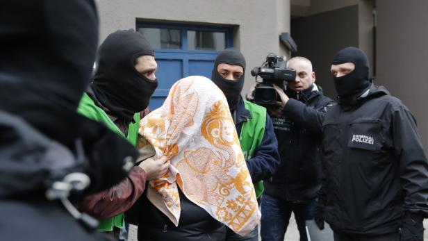 Vermummte Personen, darunter ein Beamter der Kriminalpolizei, führen eine Person mit einem Handtuch über dem Kopf ab.