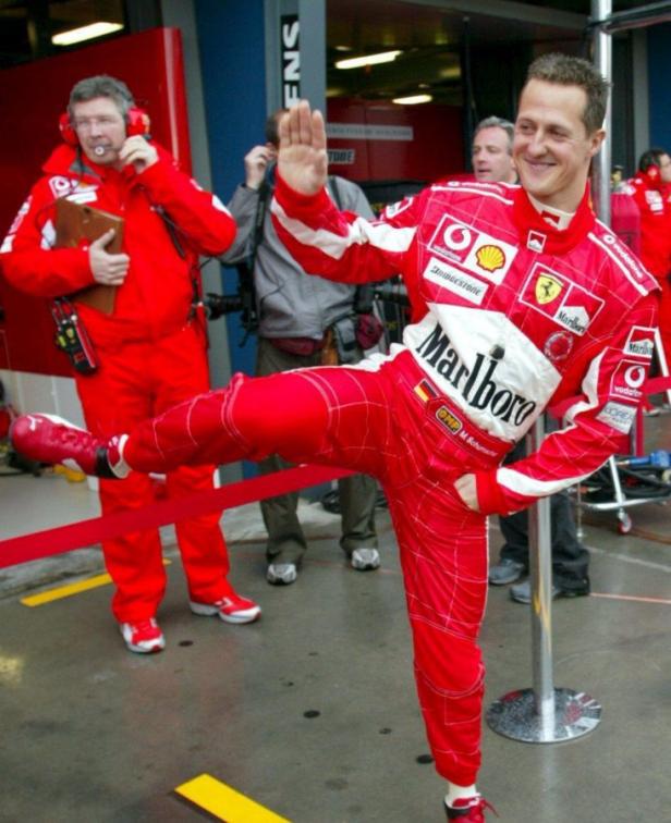 Michael Schumacher posiert in rotem Rennfahrer-Overall mit erhobenem Bein.