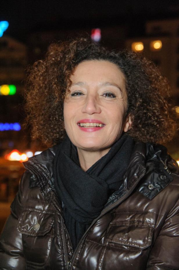 Eine Frau mit lockigem Haar lächelt in die Kamera, bekleidet mit einer braunen Jacke und einem schwarzen Schal.