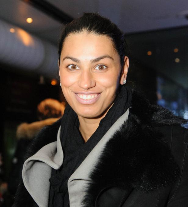 Eine lächelnde Frau mit dunklen Haaren und einem schwarzen Mantel mit Pelzkragen.