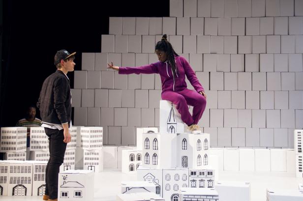 Eine Frau in lila Trainingsanzug sitzt auf einem Turm aus bemalten Kartons, während ein Mann vor ihr steht.