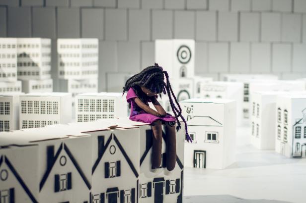 Eine Puppe mit Zöpfen sitzt auf einem Modell einer Stadt aus weißen Häusern.