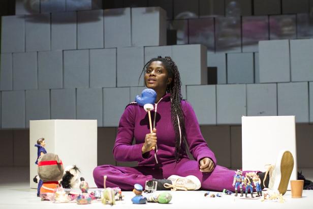 Eine Frau sitzt mit gekreuzten Beinen auf dem Boden, umgeben von Spielzeug und hält einen Boxhandschuh am Stiel.