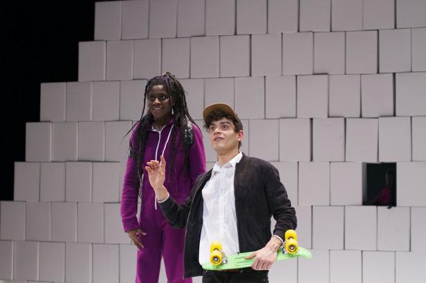 Zwei Schauspieler stehen vor einer Wand aus weißen Blöcken, einer hält ein Skateboard.