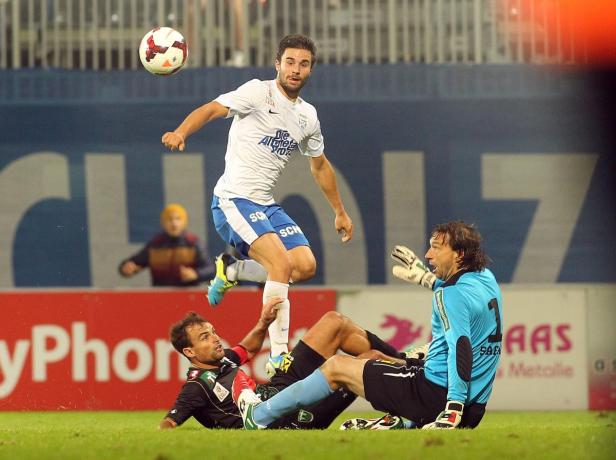 Ein Fußballspieler springt, um den Ball zu erreichen, während ein anderer am Boden liegt und der Torwart zusieht.