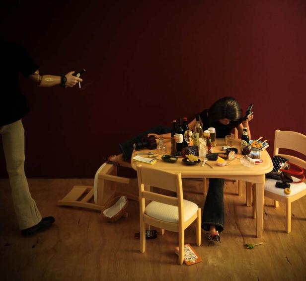 Eine Frau liegt betrunken auf einem Tisch voller leerer Flaschen, während eine Puppe mit einer Pistole auf sie zielt.