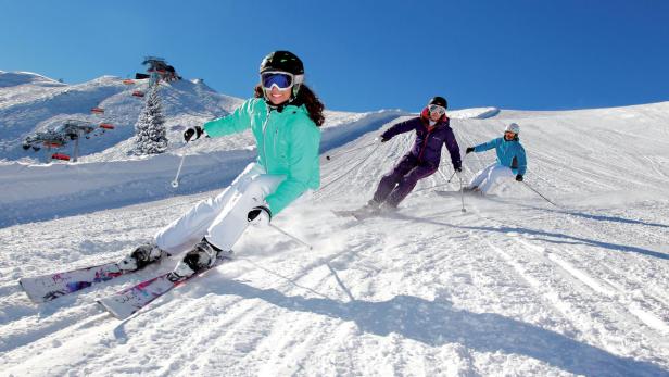 Drei Skifahrer fahren an einem sonnigen Tag eine schneebedeckte Piste hinunter.