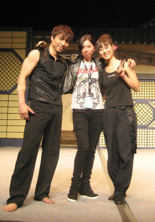 Drei asiatische Darsteller posieren auf einer Bühne vor einem japanisch anmutenden Hintergrund.