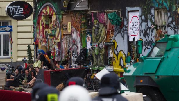 Eine Gruppe von Menschen sitzt vor einem mit Graffiti bedeckten Gebäude, während ein gepanzertes Fahrzeug im Vordergrund steht.