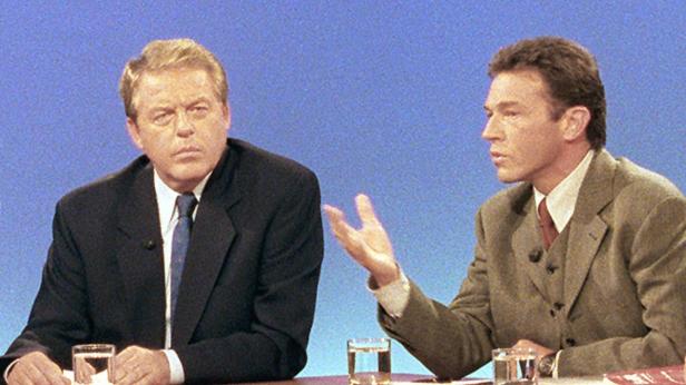 Zwei Männer sitzen an einem Tisch während einer Fernsehdiskussion.