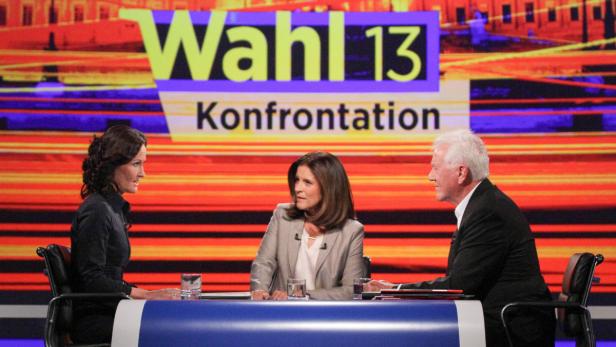 Drei Personen sitzen an einem Tisch vor dem Hintergrund der „Wahl 13“-Konfrontation.