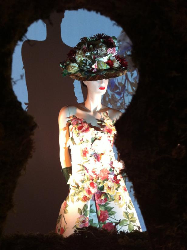 Eine Schaufensterpuppe trägt ein florales Kleid und einen Hut, gesehen durch einen dunklen Durchgang.