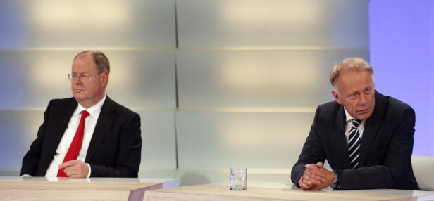 Zwei Männer in Anzügen sitzen an einem Tisch vor einer neutralen Wand.