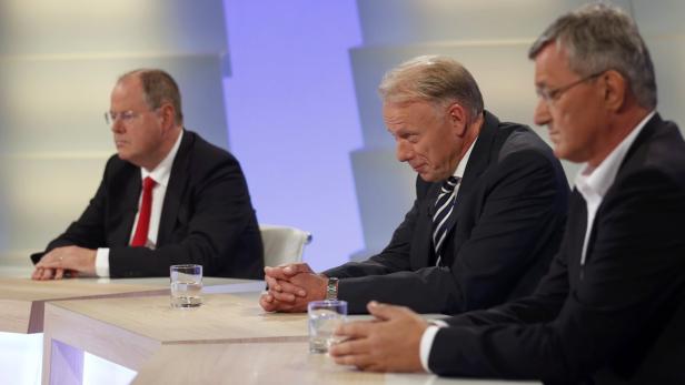 Drei Männer in Anzügen sitzen an einem Tisch mit Wassergläsern.