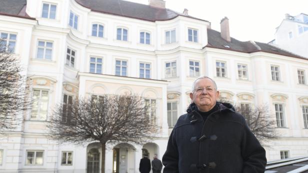 Ein älterer Mann mit Brille steht vor einem hellen, mehrstöckigen Gebäude.