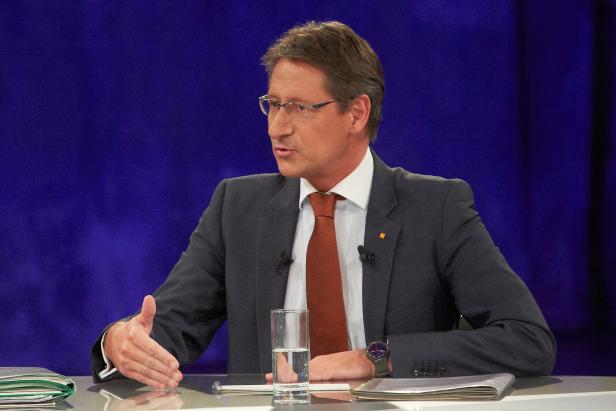 Ein Mann mit Brille und Anzug gestikuliert an einem Tisch mit Wasserglas.