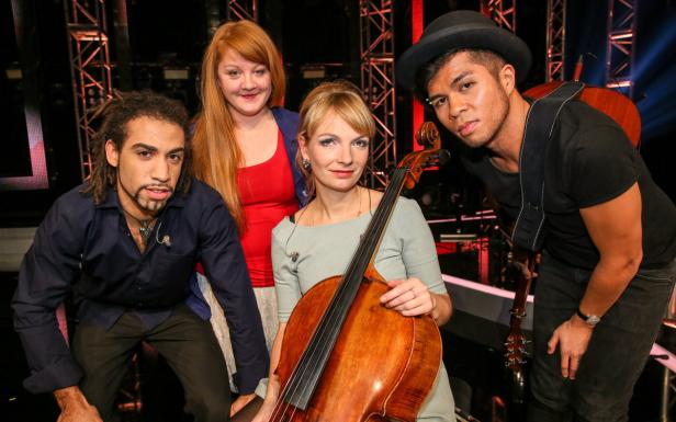 Eine Musikgruppe posiert mit einem Cello vor einem dunklen Hintergrund.