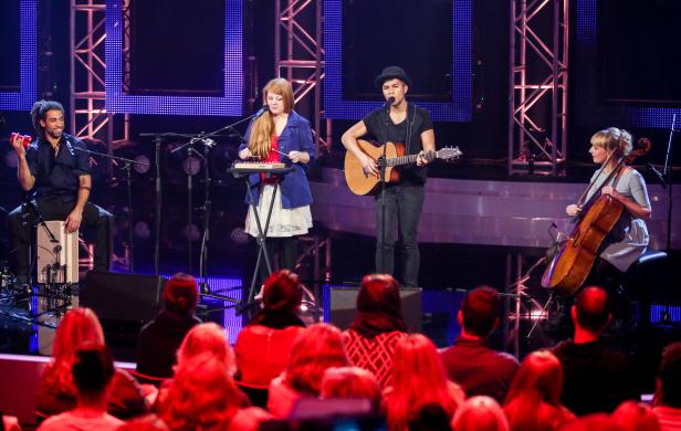 Eine Band spielt auf einer Bühne vor Publikum mit Gitarre, Cello, Xylophon und Perkussion.