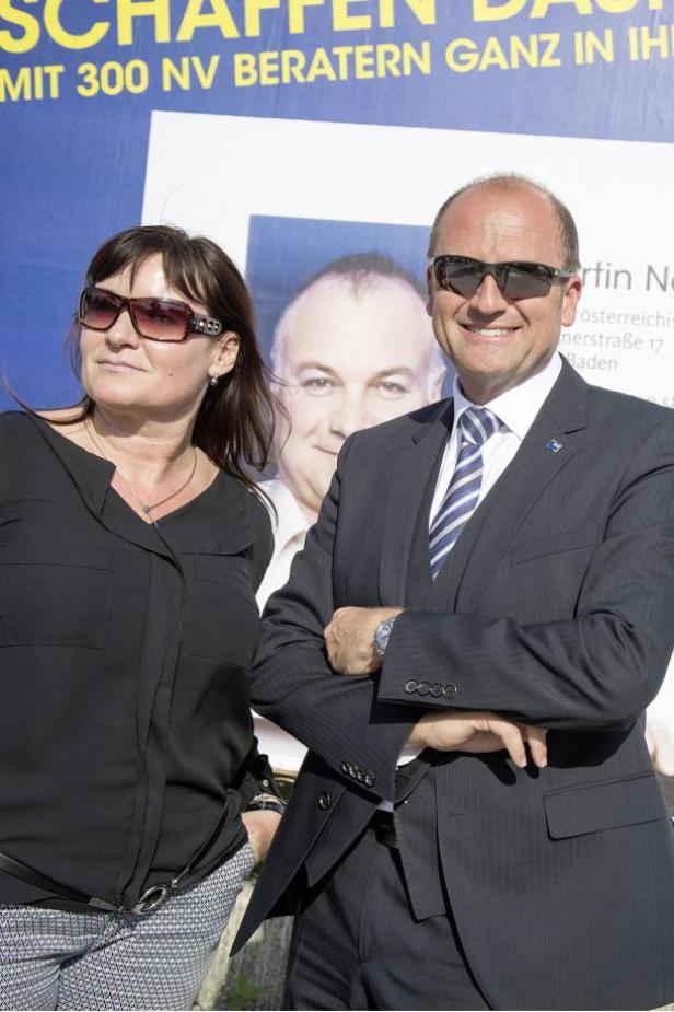 Ein Mann im Anzug und eine Frau mit Sonnenbrille stehen vor einem Wahlplakat.