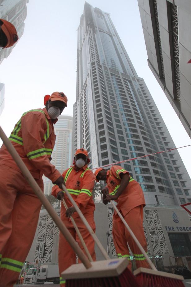 Drei Straßenkehrer reinigen mit Besen eine Straße in Dubai vor dem Hochhaus The Torch.