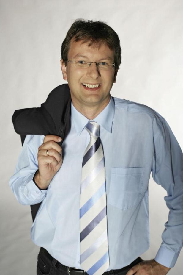 Ein lächelnder Mann mit Brille, Hemd und Krawatte, mit Jackett über der Schulter.