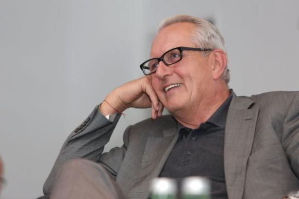 Ein älterer Mann mit Brille und grauem Anzug lächelt und ruht seinen Kopf auf seiner Hand.