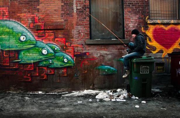 Ein Mann sitzt auf einer Mülltonne und angelt vor einer mit Graffiti bemalten Wand.