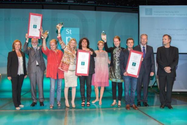Angelika Sery-Froschauer (WKÖ; ganz links), Reinhold Mitterlehner und Jurypräsident Paul Harather (ganz rechts) nehmen die Werbekampagnen-Sieger von Verbund, TBWA\Wien und Jerk Films in die Mitte. Eine Gruppe von Personen posiert mit Urkunden und Preisen auf einer Bühne.