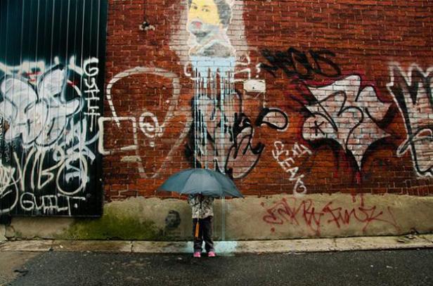 Ein Kind mit Regenschirm steht vor einer mit Graffiti bedeckten Ziegelmauer.