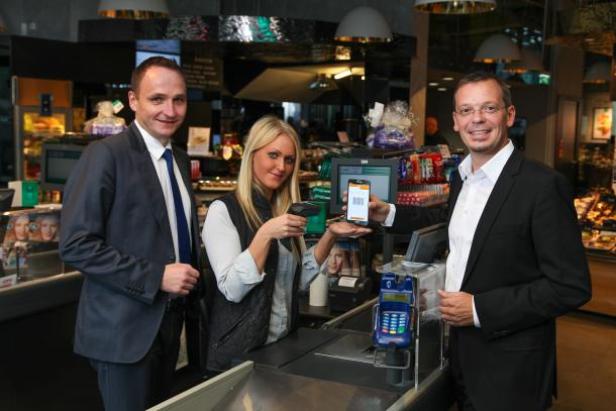 Zwei Männer in Anzügen und eine Frau stehen an einer Supermarktkasse.