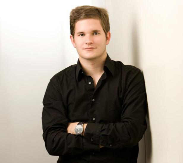 Ein junger Mann mit verschränkten Armen lehnt an einer weißen Wand.