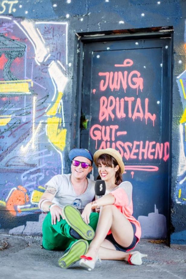 Ein Mann mit Sonnenbrille und eine Frau mit Mikrofon sitzen vor einer Wand mit Graffiti, auf der „Jung, brutal, gutaussehend“ steht.