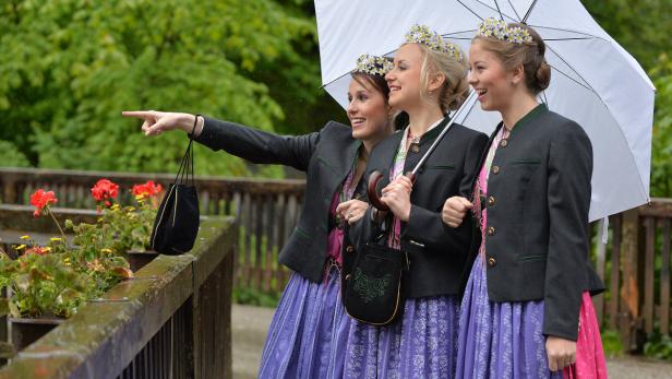 Drei junge Frauen in Tracht stehen im Regen unter einem Schirm und schauen in die Ferne.