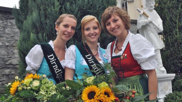 Drei junge Frauen in Dirndln, möglicherweise Dirndlprinzessinnen, posieren mit Blumensträußen vor einer Statue.