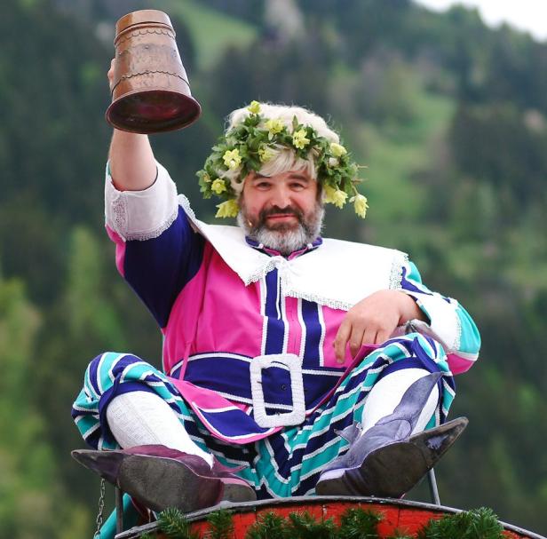 Ein Mann in Tracht mit Blumenkranz hält einen Bierkrug in die Höhe.