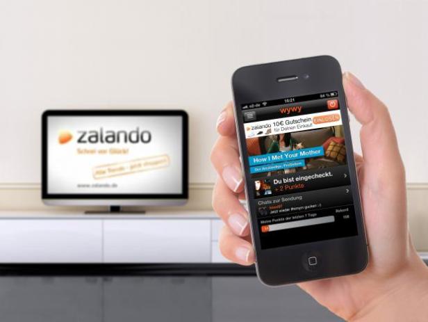 Eine Hand hält ein Smartphone mit der geöffneten Zalando-App; im Hintergrund ein Fernseher mit dem Zalando-Logo.