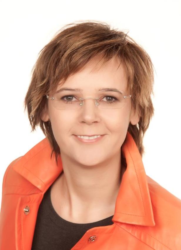 Eine Frau mit Brille und kurzem braunen Haar trägt eine orangefarbene Jacke.