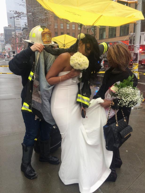 Eine Braut wird von Feuerwehrleuten unter einem gelben Regenschirm beschützt.