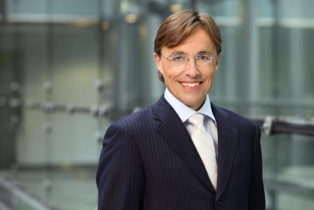 Ein lächelnder Mann mit Brille und Anzug vor einem modernen Gebäude.