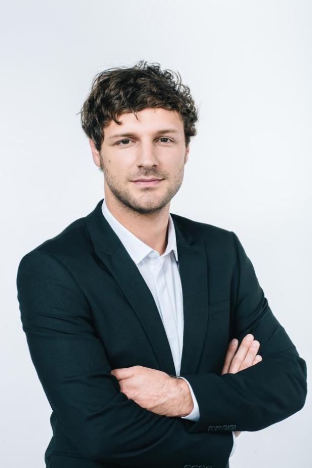 Jan Gorfer, Director Communication Consultant Carat, stellt hier mit Ein Mann mit dunklen Locken und Anzug steht mit verschränkten Armen vor einem weißen Hintergrund.