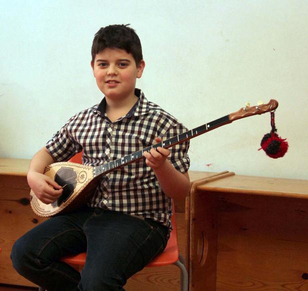 Ein Junge sitzt auf einem Stuhl und spielt ein Saiteninstrument, möglicherweise eine Bouzouki.